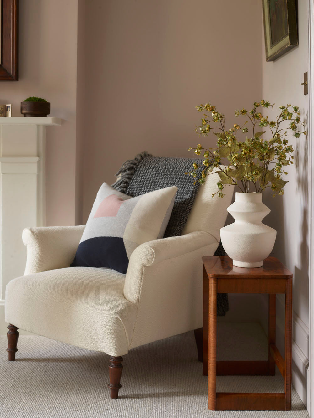 A cozy cream armchair with a patterned cushion and gray throw sits beside a wooden side table topped with a white vase holding Faux Winter Cherry, Green - 92 cm stems in a softly lit, neutral-toned living room.